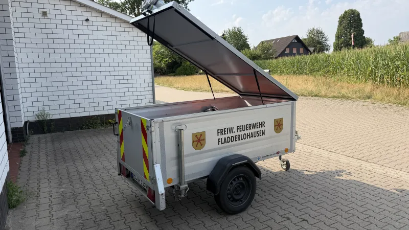 Silberner Materialanhänger mit Aufschrift Freiw. Feuerwehr Fladderlohausen und aufgeklappter Haube, neben dem Gerätehaus abgestellt.