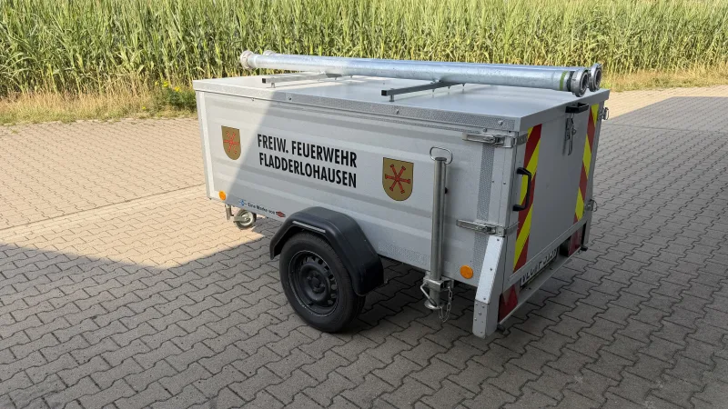 Der geschlossene Materialanhänger von schräg hinten auf dem gepflasterten Vorplatz, mit zwei Saugschläuchen auf dem Deckel.