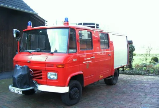 Rotes Löschgruppenfahrzeug Mercedes-Benz 408 mit Ziegler-Aufbau und Blaulichtern auf einem gepflasterten Hof vor einem dunklen Holzschuppen.