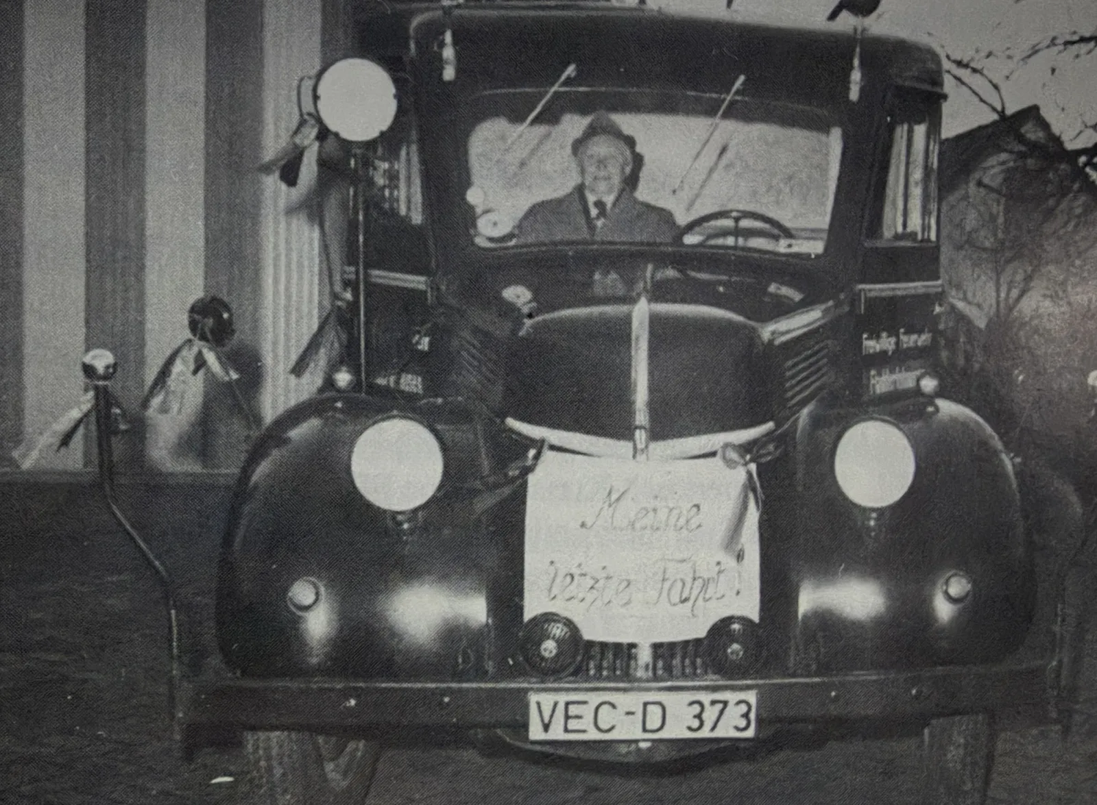Schwarz-Weiß-Foto des alten LF 8 von vorn mit dem Kennzeichen VEC-D 373 und einem Schild „Meine letzte Fahrt!“ am Kühler; am Steuer ein älterer Mann mit Hut.