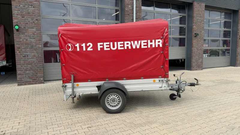 Einachsiger Materialanhänger mit roter Plane und Aufschrift 112 Feuerwehr vor den Toren des Feuerwehrhauses.