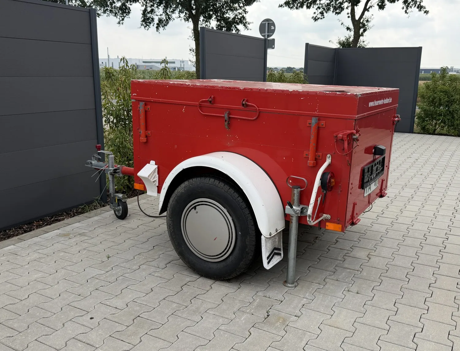 Kleiner roter Anhänger mit geschlossenem Kastenaufbau auf der gepflasterten Terrasse am Feuerwehrhaus.