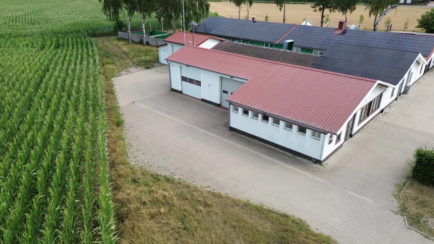 Luftbild des Feuerwehrhauses Fladderlohausen: weisses Gebäude mit rotem Blechdach und Toren zum gepflasterten Vorplatz, dahinter ein angrenzendes langes Gebäude mit Solarmodulen auf dem Dach, links ein Maisfeld.