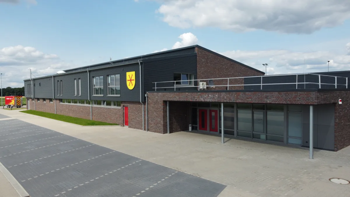 Eingangsseite des Feuerwehrhauses Holdorf mit Klinkerfassade, roter Doppeltür unter dem Vordach und einem gelben Wappen an der dunklen Metallfassade.
