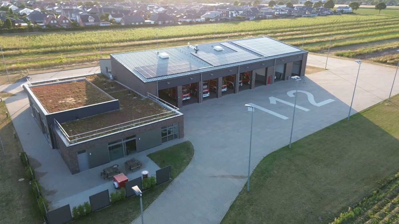 Luftbild des Feuerwehrhauses Holdorf: Fahrzeughalle mit offenen Toren und Solarmodulen auf dem Dach, davor der gepflasterte Hof mit der Bodenmarkierung 112, im Hintergrund Felder und Wohnhäuser.