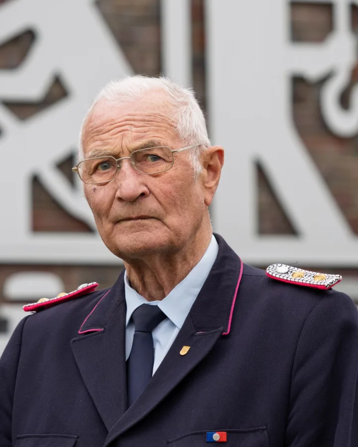 Porträt von Stefan Borgmann mit Brille in dunkelblauer Uniform vor einem großen Feuerwehrsignet aus Metall.