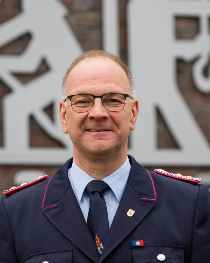 Porträt von Klaus Meyer mit Brille in dunkelblauer Uniform vor einem großen Feuerwehrsignet aus Metall.