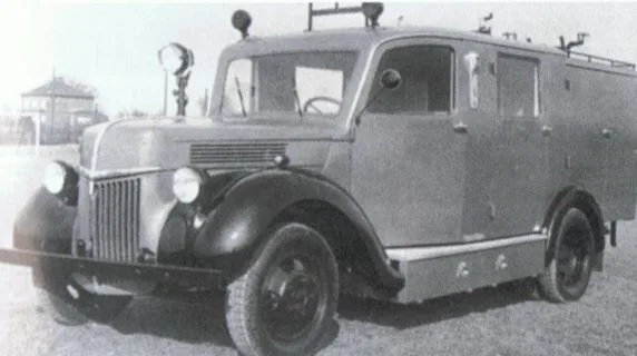 Schwarz-Weiß-Foto eines Löschgruppenfahrzeugs Ford Ruhr mit runden Kotflügeln, Suchscheinwerfer und Leiter auf dem Dach.