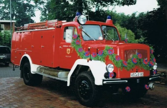 Rotes Tanklöschfahrzeug von Magirus mit weißen Kotflügeln, geschmückt mit einer Blumengirlande, auf einem gepflasterten Platz.