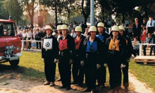 Die Wettkampfgruppe in dunklen Uniformen, Helmen und Wettkampfleibchen steht in einer Reihe auf einem Wettkampfplatz, dahinter Zuschauer und ein Löschfahrzeug.