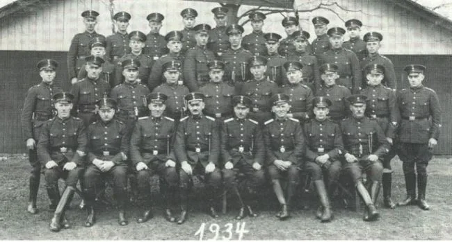Schwarz-Weiß-Gruppenfoto von 1934: Feuerwehrmänner in Uniform und Mütze in mehreren Reihen vor einem Holzgebäude, die vordere Reihe sitzend.