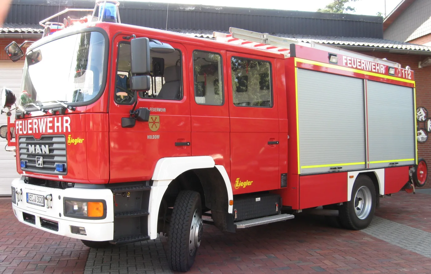 Rotes Löschgruppenfahrzeug von MAN mit Ziegler-Aufbau und Rollläden am Geräteraum, vor dem Feuerwehrhaus.