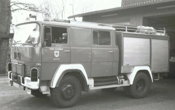 Schwarz-Weiß-Foto eines Löschgruppenfahrzeugs von Magirus mit der Aufschrift Freiw. Feuerwehr Holdorf, vor einem Gebäude geparkt.