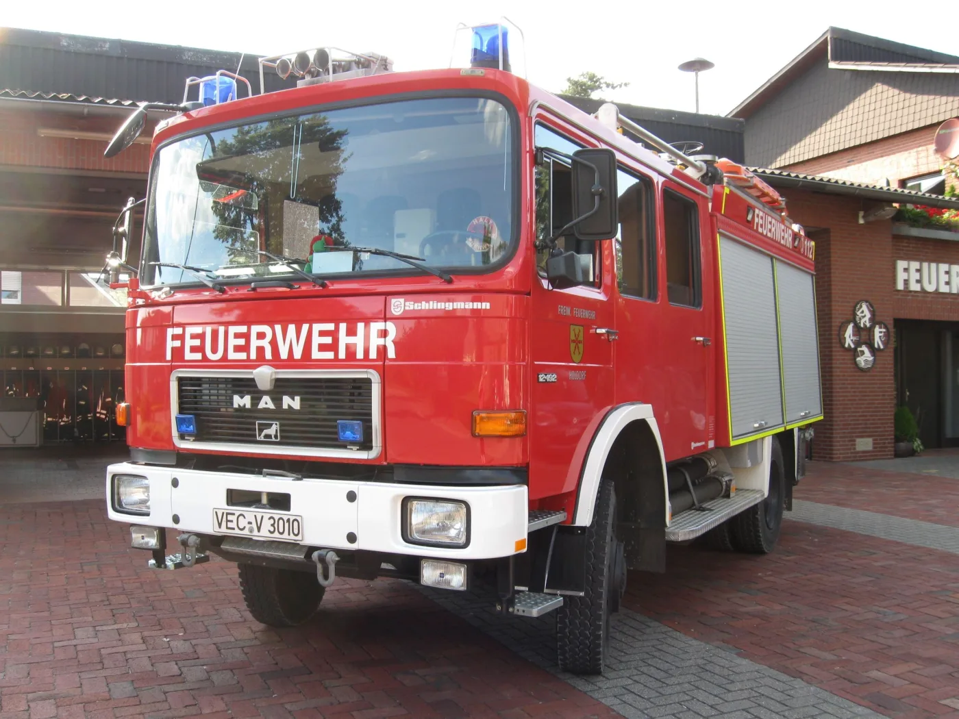 Rotes Tanklöschfahrzeug von MAN mit Schlingmann-Aufbau, frontal vor dem alten Feuerwehrhaus aus Klinker.