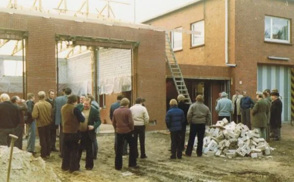 Baustelle am Feuerwehrhaus: offener Rohbau mit Dachstuhl, davor stehen Gruppen von Männern, rechts ein Stapel Steine.