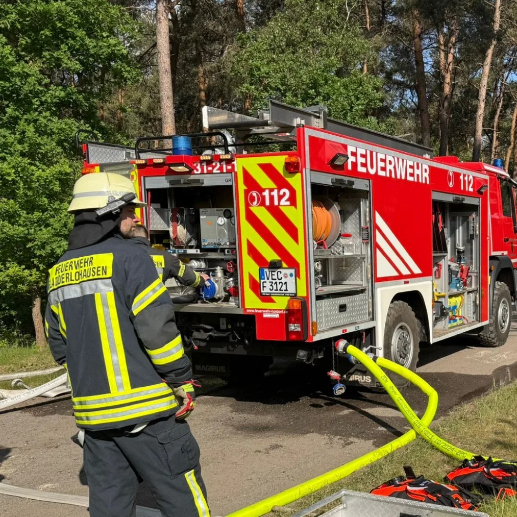 Eine Einsatzkraft der Feuerwehr Fladderlohausen steht am geöffneten Heck eines Löschfahrzeugs im Wald, an das ein gelber Schlauch angeschlossen ist.