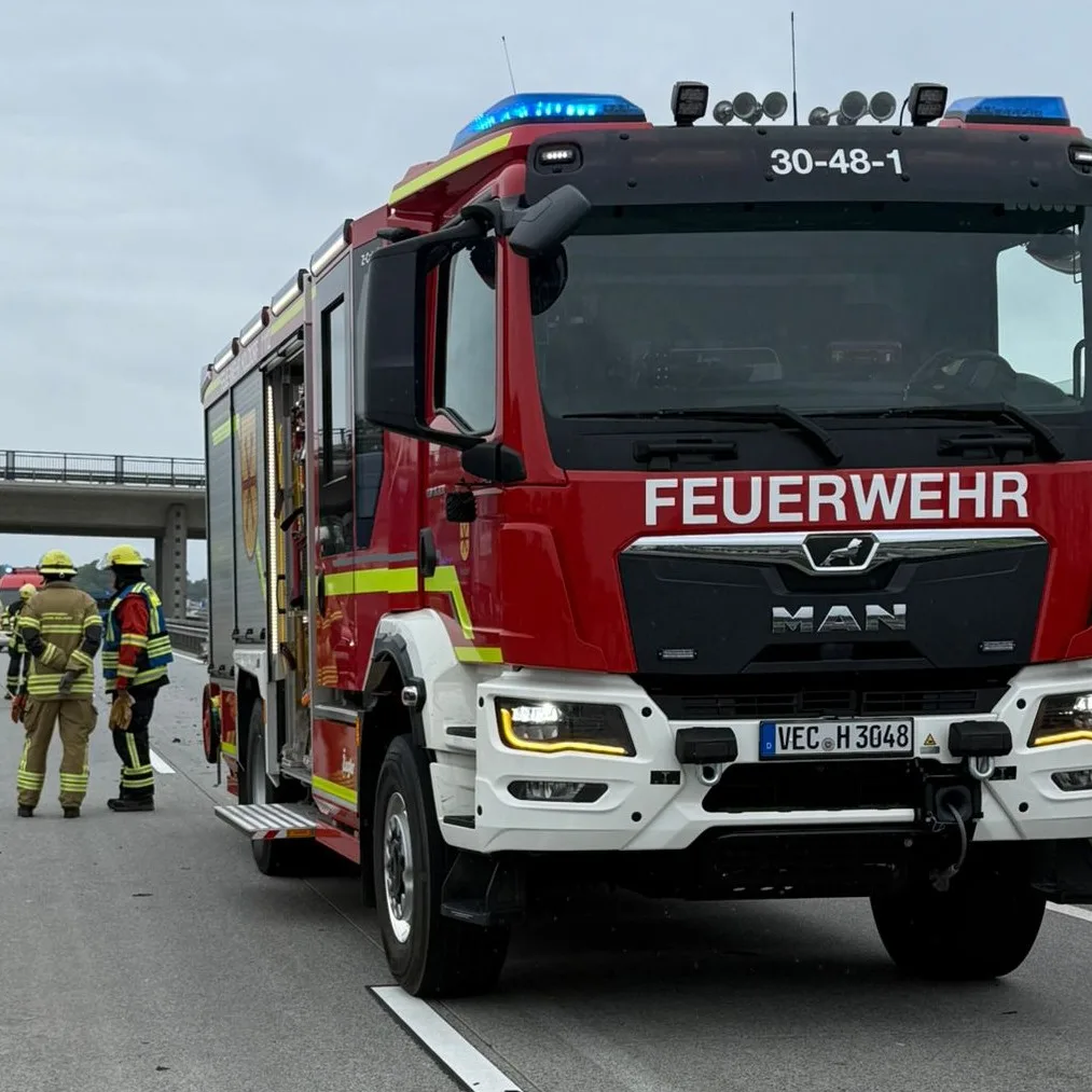 Ein Löschfahrzeug der Feuerwehr Holdorf mit dem Funkrufnamen 30-48-1 steht mit Blaulicht auf einer Autobahn, links daneben zwei Einsatzkräfte, im Hintergrund eine Brücke.