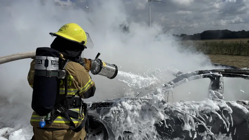 Eine Einsatzkraft der Feuerwehr Holdorf mit Atemschutzgerät löscht ein ausgebranntes Auto mit Schaum, von hinten gesehen.