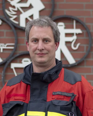 Porträt von Stefan Lüpke-Narberhaus in roter Einsatzjacke vor einer Klinkerwand mit Feuerwehrsignet.